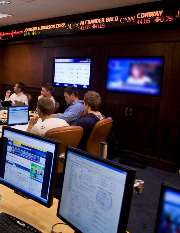students sitting at conference table behind monitors showing stock prices