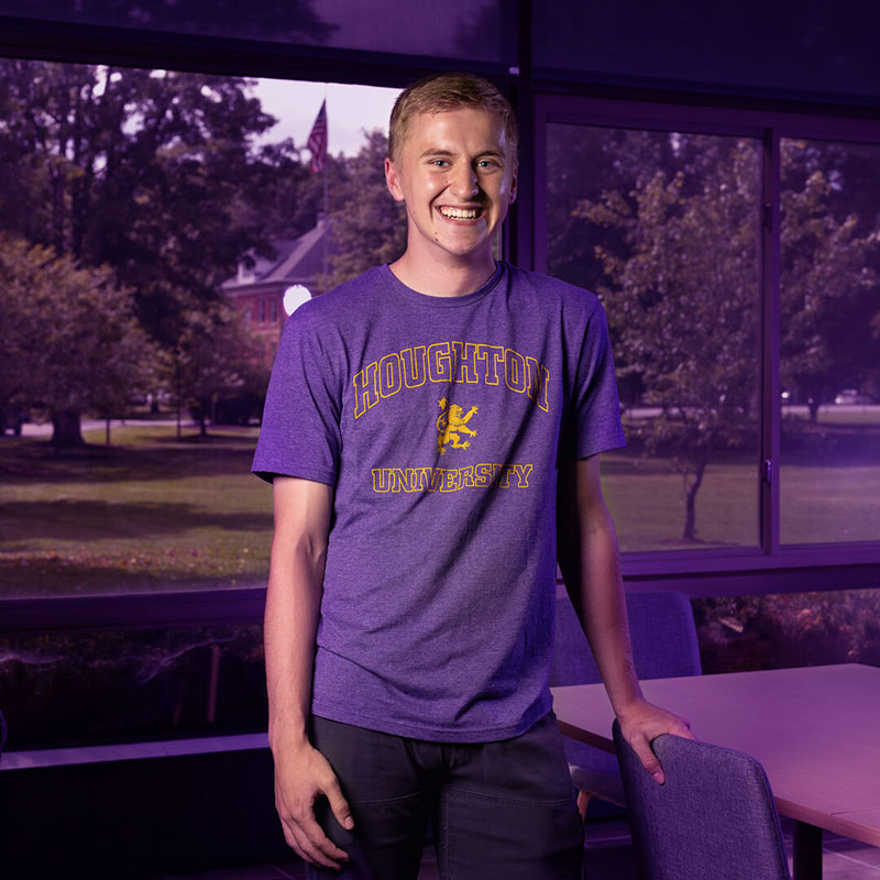 Houghton student Ryan Kullander wearing a purple Houghton shirt standing in conference room with view of campus in the back.